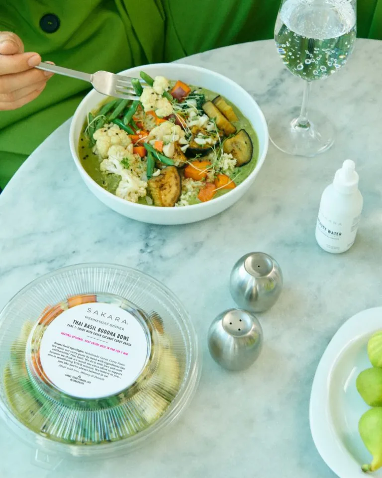 Person in a bright green blazer eating a bowl of Sakara curry at a marble table with a glass of sparkling water.