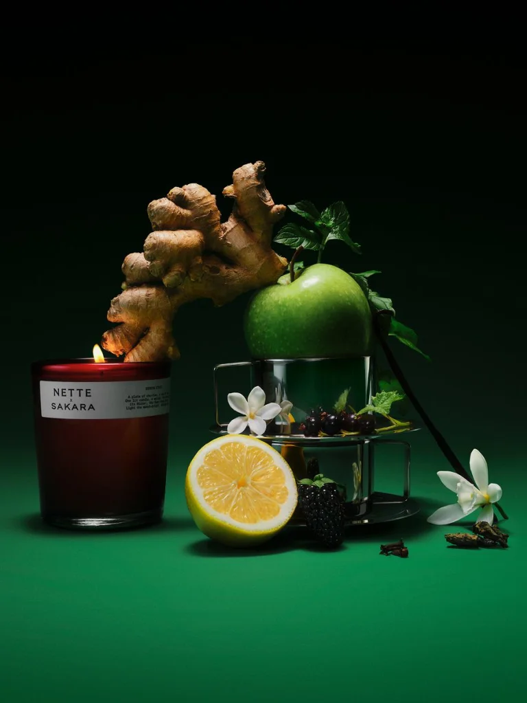 Sakara x Nette Serene State Scented Candle in deep red translucent glass vessel with white label, shown lit alongside green apple, lemon, ginger, berries, jasmine, mint, and clove representing fragrance notes.