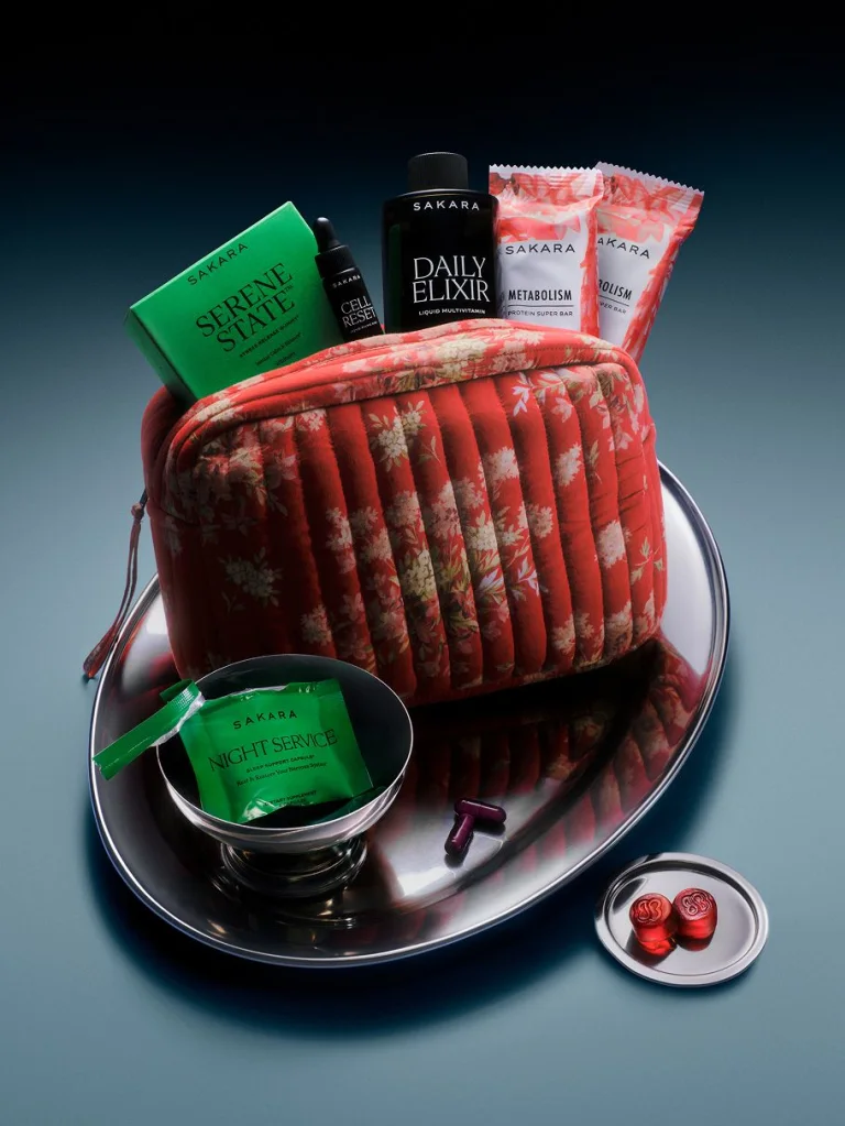 Sakara x DÔEN Holloway Pouch in red quilted floral cotton with zipper closure, shown filled with Daily Elixir deluxe sample 3.25 fl oz, Cell Reset deluxe sample 0.17 fl oz, Serene State 3 packs of 2 gummies, Night Service deluxe sample 1 pack of 6 capsules, and 2 Metabolism Protein Super Bars 45g each.