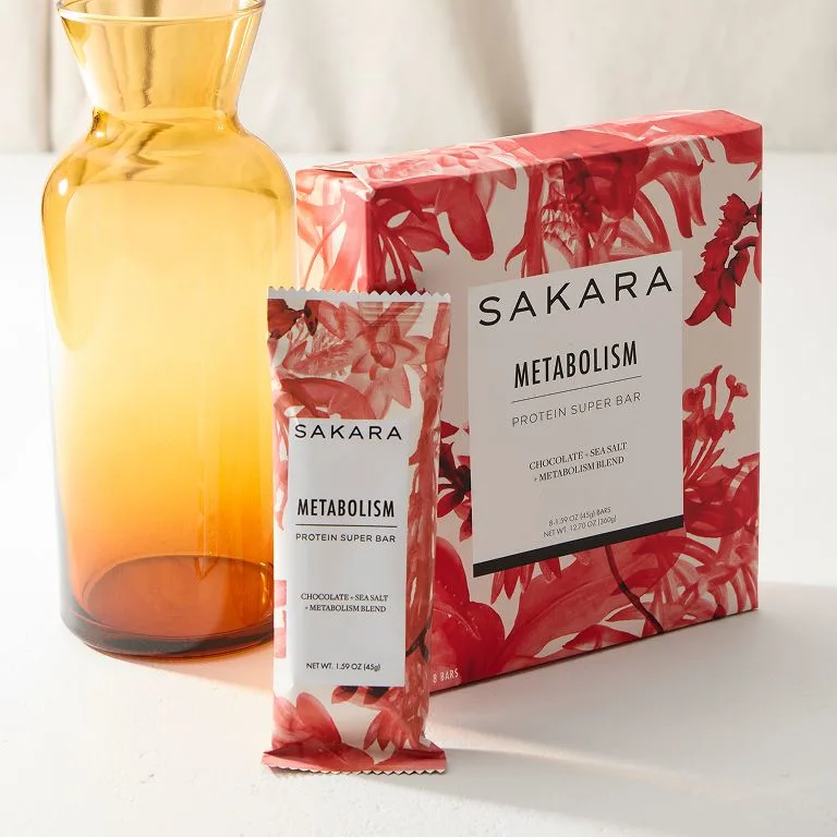 Sakara Metabolism Super Bar box and wrapped bar displayed beside an amber glass carafe on a light background.