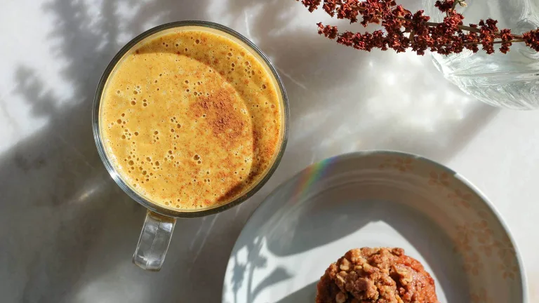Frothy golden latte in a glass mug beside a muffin on a plate.