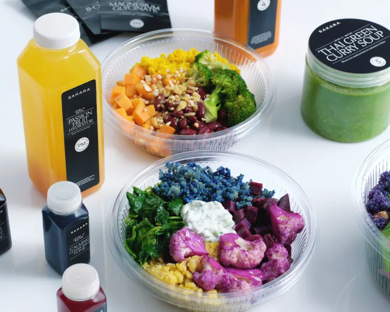 Sakara Level II: Detox ready-to-eat meals in clear containers featuring broccoli, beans, grains, pink cauliflower, and leafy greens displayed with bottled Passion Fruit Hydrator and Calming Chamomile Tonic.
