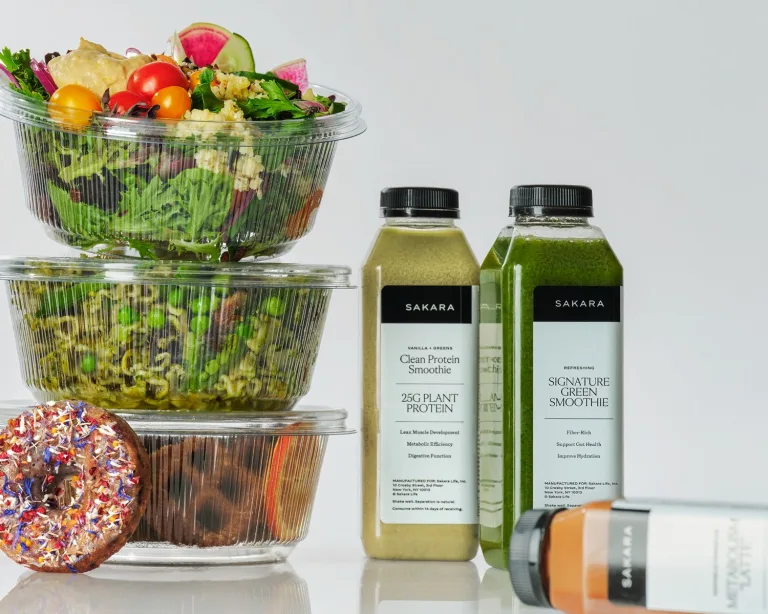 Sakara Signature Nutrition Program assortment showing clear plastic ready-to-eat meal containers, a sprinkle-topped donut, and bottled smoothies labeled “Clean Protein Smoothie – 25G Plant Protein” and “Signature Green Smoothie.”