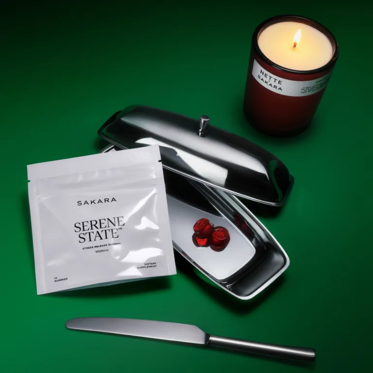 Serene State stress support gummies in a white pouch beside a lit Nette candle and a silver tray with red gummies on a green background.