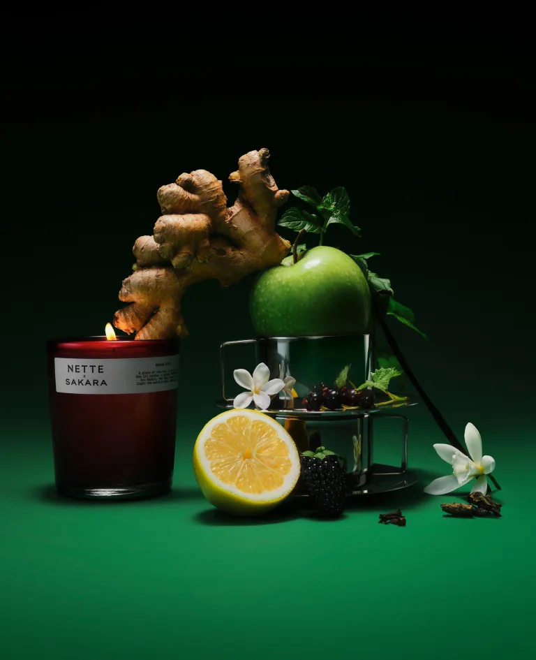 Sakara x Nette Serene State Scented Candle in deep red translucent glass vessel with white label, shown lit alongside green apple, lemon, ginger, berries, jasmine, mint, and clove representing fragrance notes.