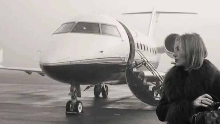 Black-and-white image of a private jet on a runway with a woman standing beside it.