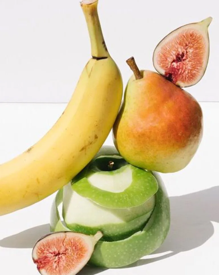 Banana balanced on stacked green apple slices with pears and figs on a white surface.