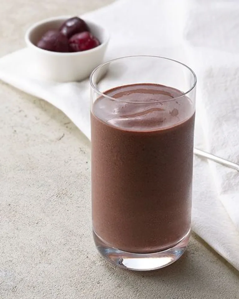 Glass of chocolate almond butter protein smoothie on a neutral surface with a white napkin and cherries in the background.