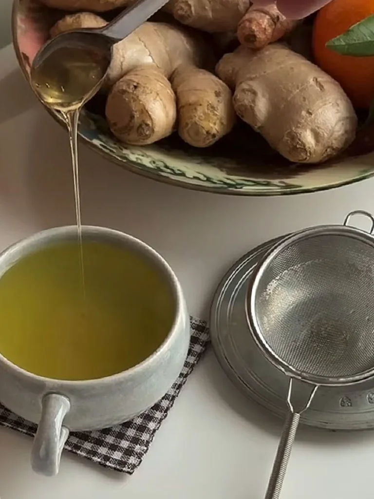 Ginger slices on a cutting board and honey being poured into a cup of tea with cinnamon and spices nearby.