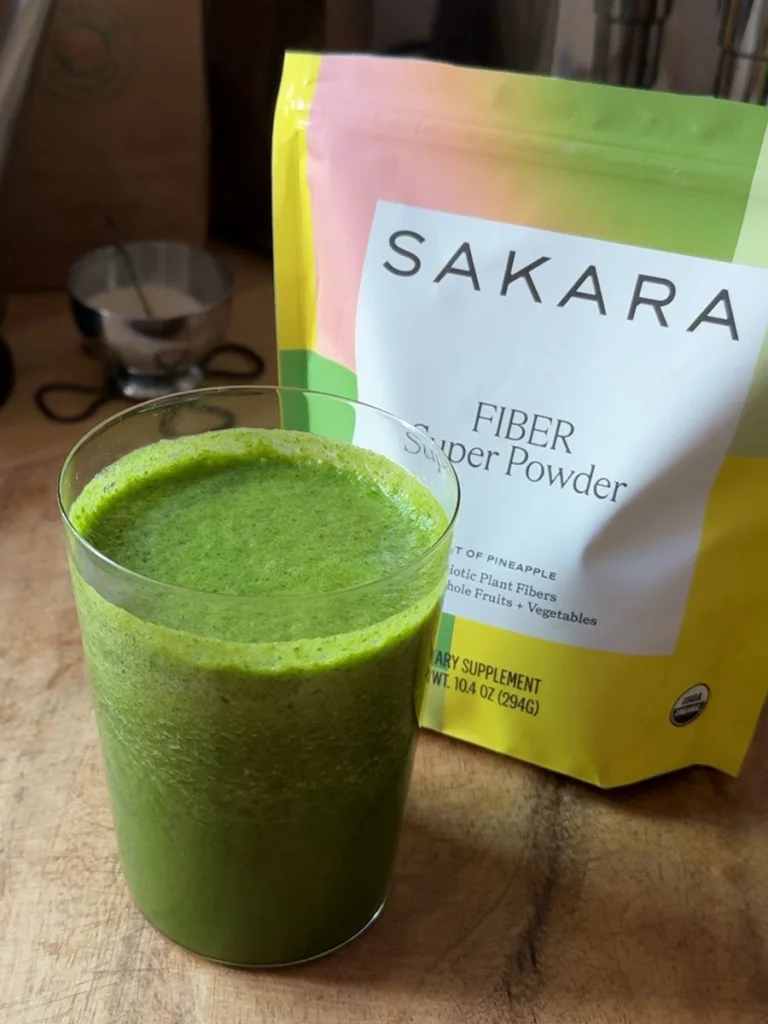 Glass of green smoothie beside Sakara Fiber Super Powder pouch, apple slices, pineapple, and greens.