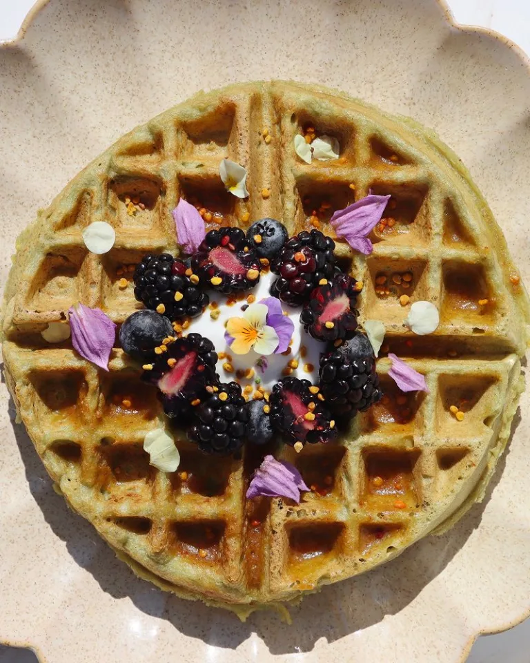 Golden waffle topped with berries, yogurt, and edible flowers on a scalloped plate.