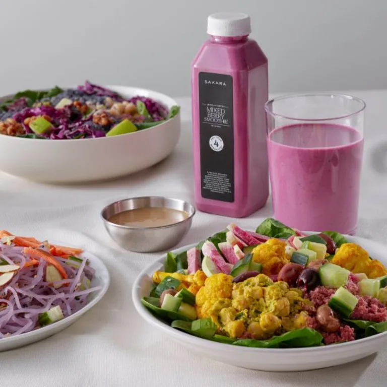 Sakara Level II: Detox meals arranged on a white table including mixed salads, a pink smoothie bottle with matching glass, and small bowls of dressing.