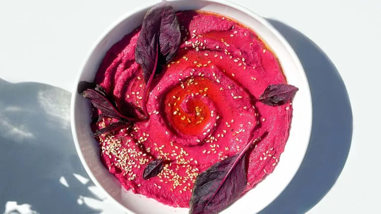White bowl filled with pink beed hummus topped with purple leafy greens and seeds.