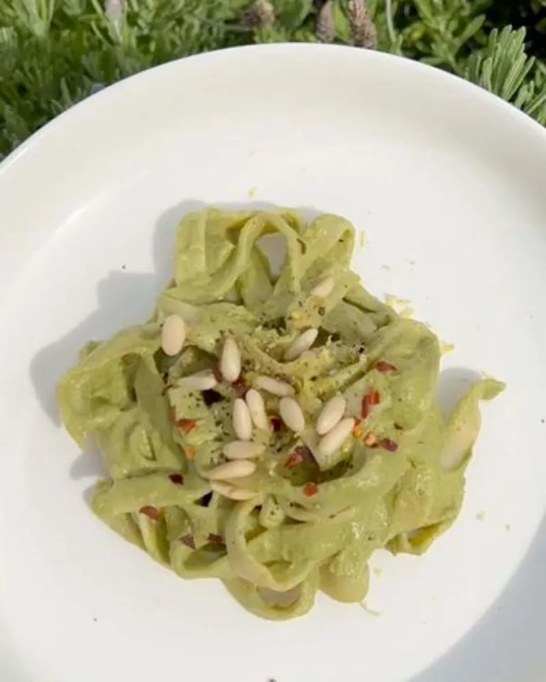 A close-up of creamy green sauce-coated noodles, topped with seeds and red pepper flakes in a white bowl.