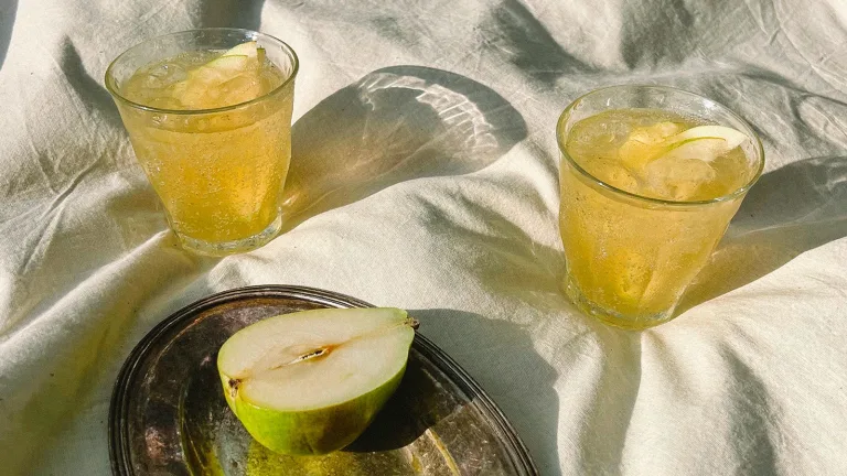 Two glasses of a pear ginger honey shrub beverage with ice beside a halved pear on a tray.
