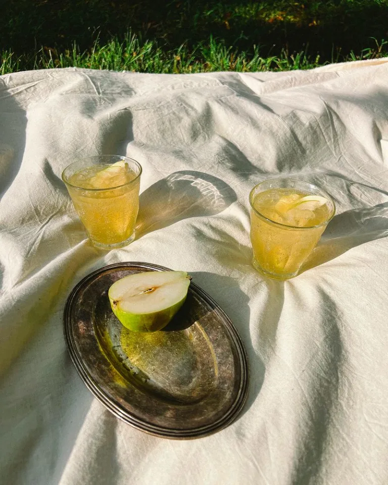 Two glasses of a pear ginger honey shrub beverage with ice beside a halved pear on a tray.