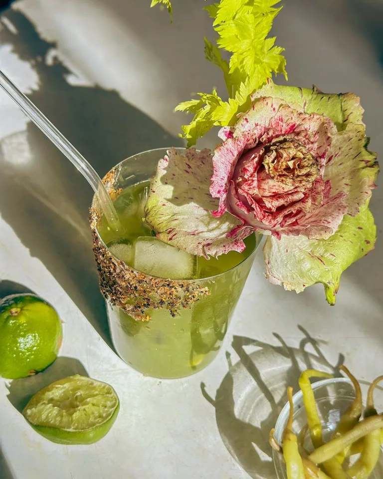 Two cocktail green juices in glasses garnished with cucumber ribbons and leafy greens.