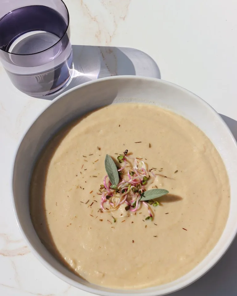 Creamy blended cauliflower soup garnished with herbs and finely sliced vegetables in a white bowl.