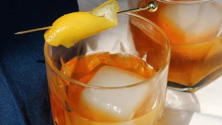 Amber-colored cocktail with large ice cubes and a lemon twist garnish in short glasses on a marble surface.