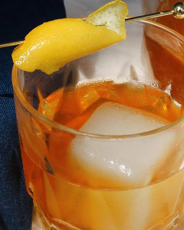 Amber-colored cocktail with large ice cubes and a lemon twist garnish in short glasses on a marble surface.