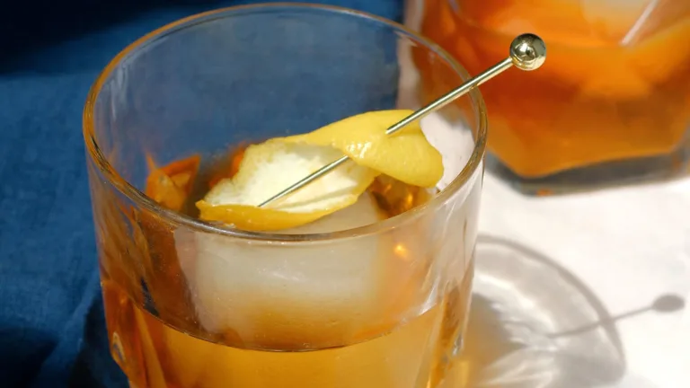 Amber-colored cocktail with large ice cubes and a lemon twist garnish in short glasses on a marble surface.