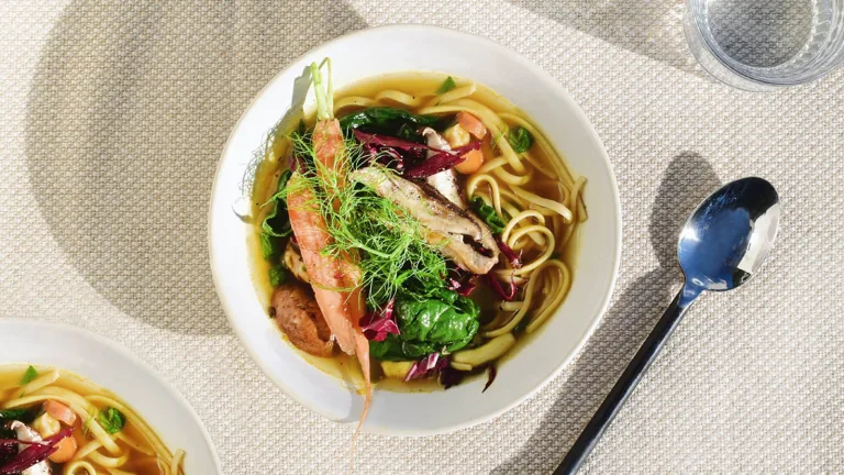 Bowl of noodle soup topped with greens and vegetables placed beside a spoon on a textured surface.