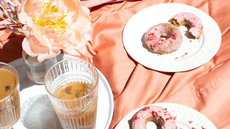 Table setting with pink-frosted donuts, iced coffee drinks, and a flower arranged on a satin fabric background.