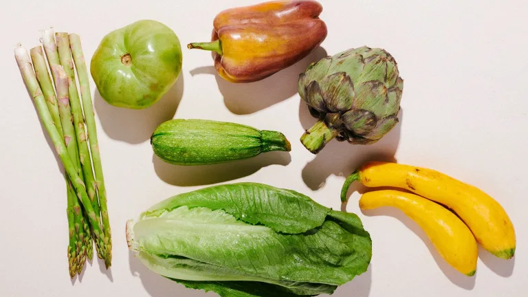 Assorted fresh produce including asparagus, green apple, bell pepper, artichoke, zucchini, romaine lettuce, and bananas arranged on a neutral surface.