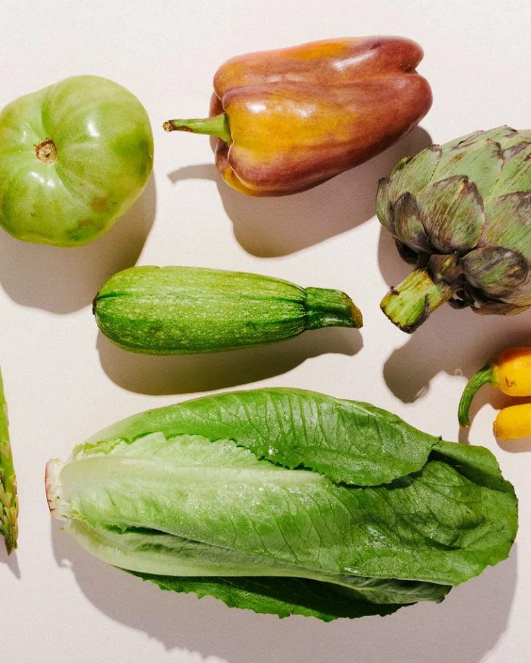 Assorted fresh produce including asparagus, green apple, bell pepper, artichoke, zucchini, romaine lettuce, and bananas arranged on a neutral surface.