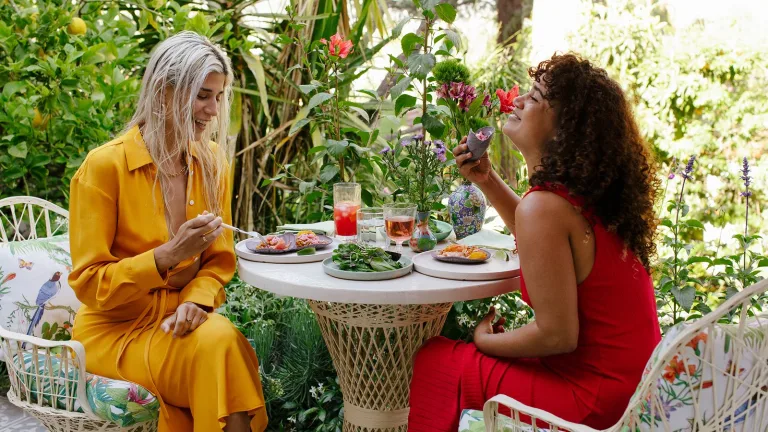 Two women sitting at an outdoor table in a garden, eating and drinking together.