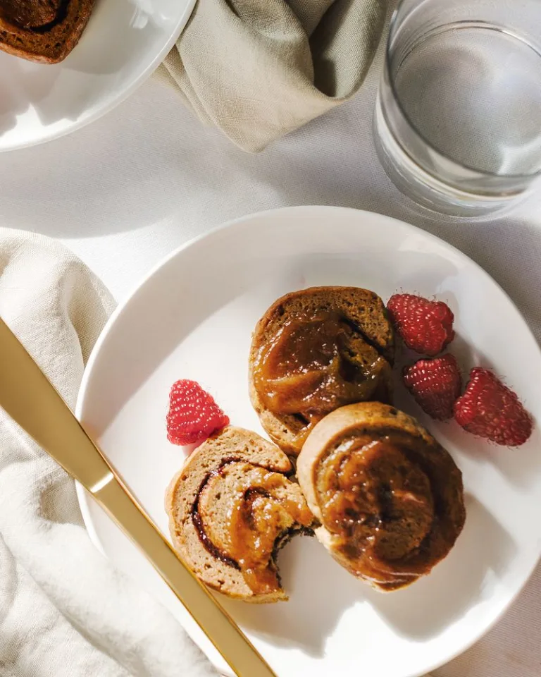 Breakfast plate with cinnamon rolls topped with caramel sauce, raspberries, peeled citrus, and a knife on a white tablecloth.