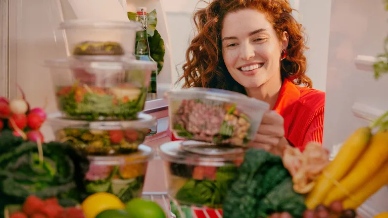 Smiling woman placing stacked Sakara meal containers into a refrigerator filled with fresh produce.