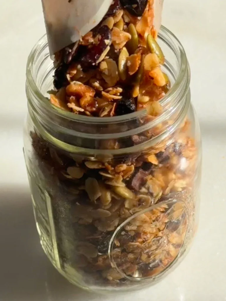 Spoon pouring crunchy granola with nuts and dried fruit into a glass jar.