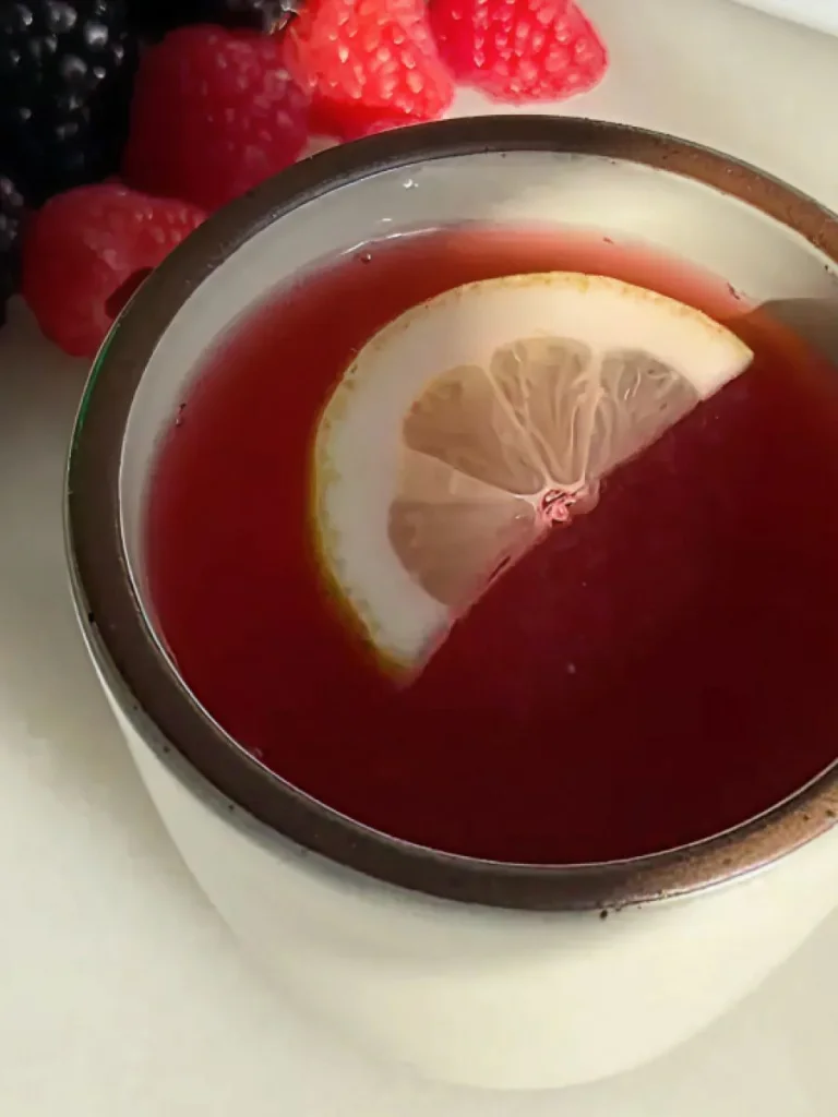 Mug of deep red cider garnished with a lemon slice, with berries in the background.