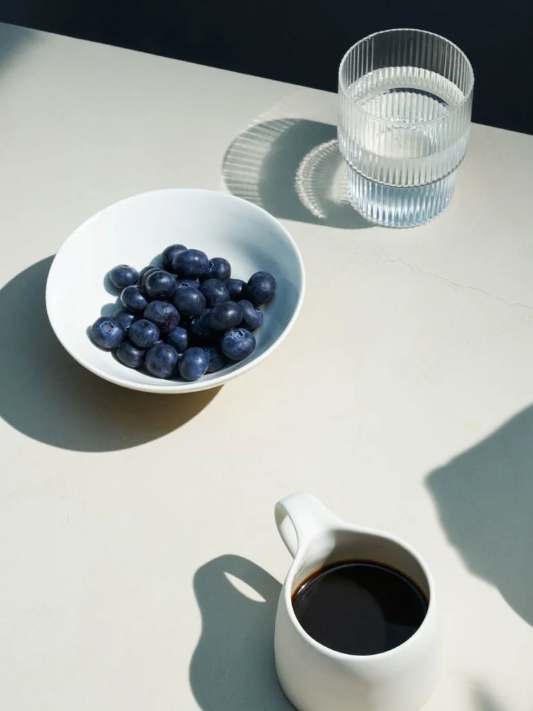 Small bowl of blueberries beside a glass of water and a cup of black coffee on a white table.
