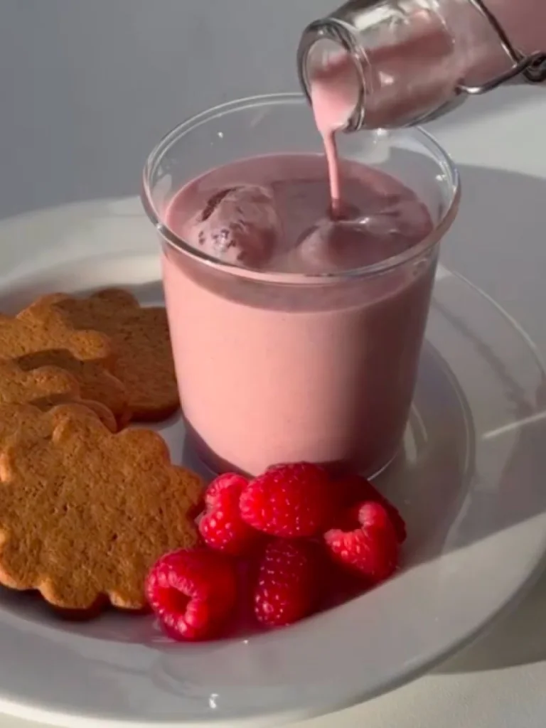 A glass of pink-colored strawberry milk on a plate with cookies and raspberries.