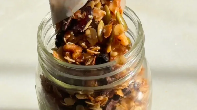 Spoon pouring crunchy granola with nuts and dried fruit into a glass jar.