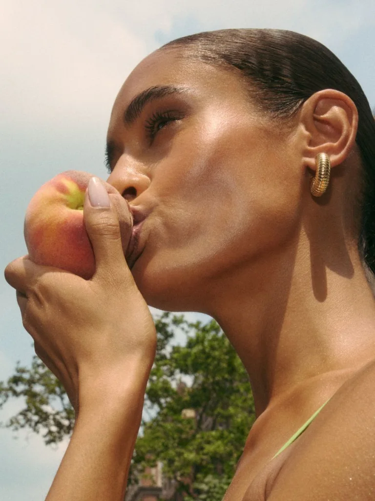 A close-up image of a woman taking a bite out of a peach, with a background of NYC.