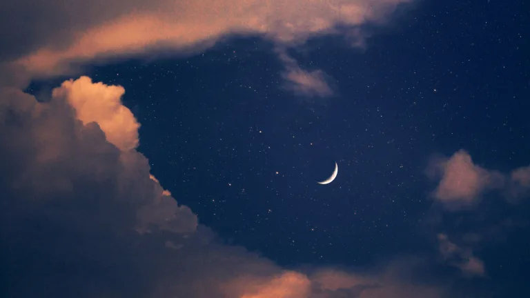 Crescent moon glowing in a deep navy night sky filled with scattered stars, framed by soft, billowing clouds tinged with warm peach and pink hues, creating a dreamy, atmospheric celestial scene.